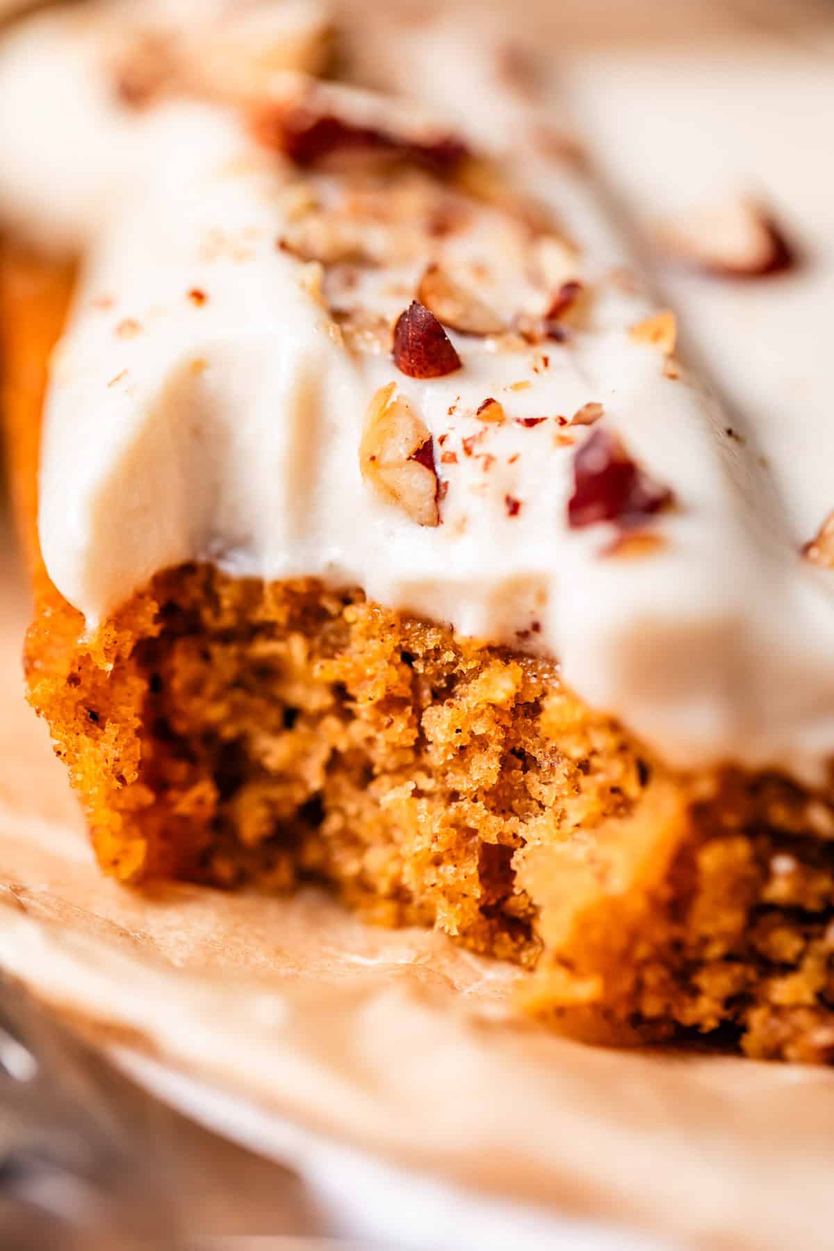 A close-up of a slice of moist, spiced cake topped with creamy white frosting and sprinkled with chopped nuts, resting on a piece of parchment paper.