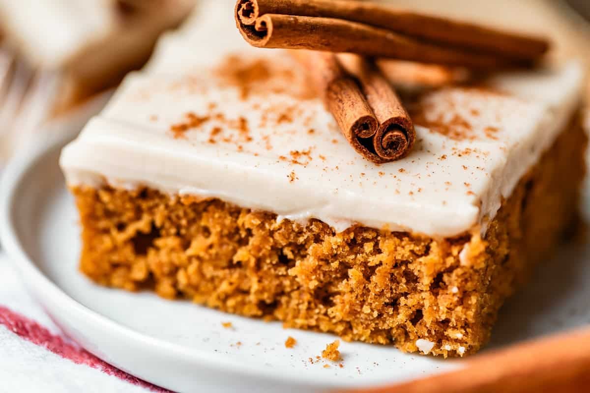 A close-up of a square piece of spiced cake with white frosting, sprinkled with cinnamon and topped with two cinnamon sticks, served on a white plate.