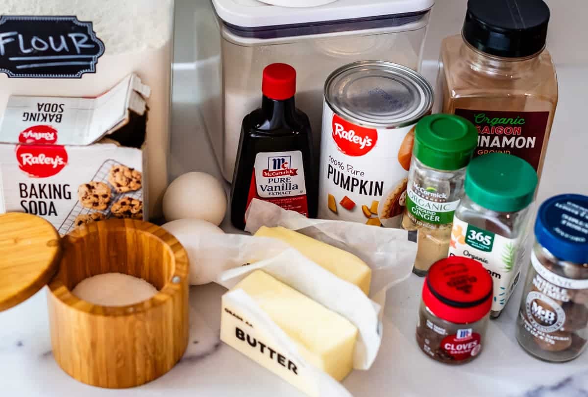A close-up of various baking ingredients on a countertop, including flour, baking soda, sugar, butter, eggs, vanilla extract, canned pumpkin, ground cinnamon, ground cloves, and ground nutmeg.