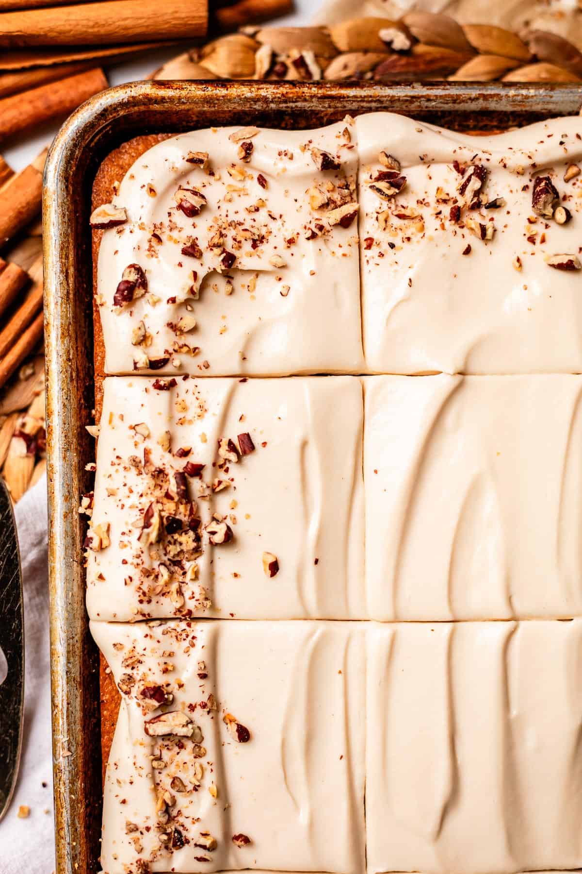 A close-up of a frosted sheet cake cut into squares, topped with chopped pecans. Swirls in the creamy frosting are visible, and cinnamon sticks surround the cake on the table.